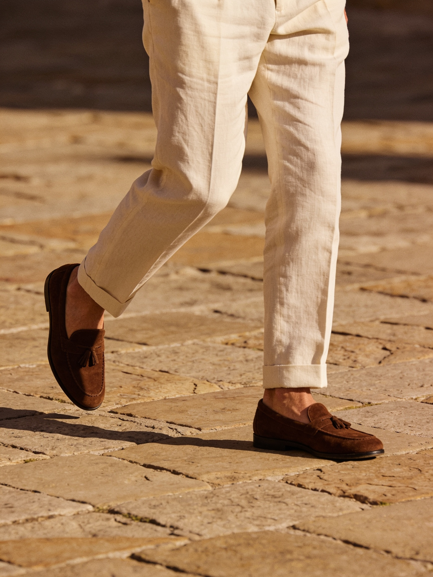 Homem caminhando em piso de pedra usando mocassim com borla em camurça marrom e calça de linho cru